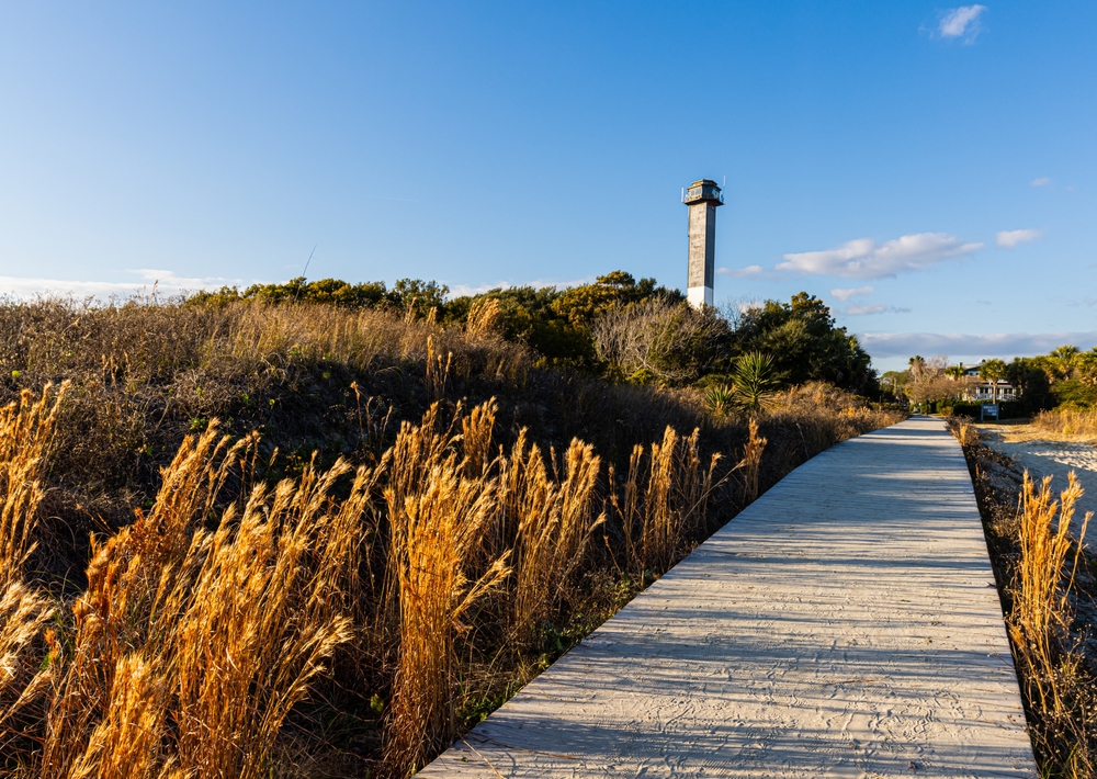 Wooden boardwalk winds through golden dune grasses toward a distant lighthouse under a clear blue sky.