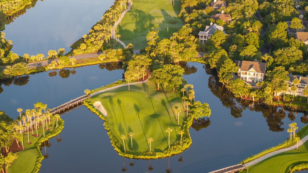 Aerial view of a golf hole on a small island, surrounded by water, with a bridge, green, and a sand bunker, near houses and trees.