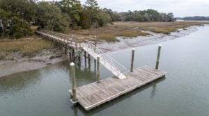 Aerial view of a residential floating dock on a Charleston tidal creek, with an aluminum gangway descending steeply from a timber walkway at low tide and the float held by timber guide piles with white caps.
