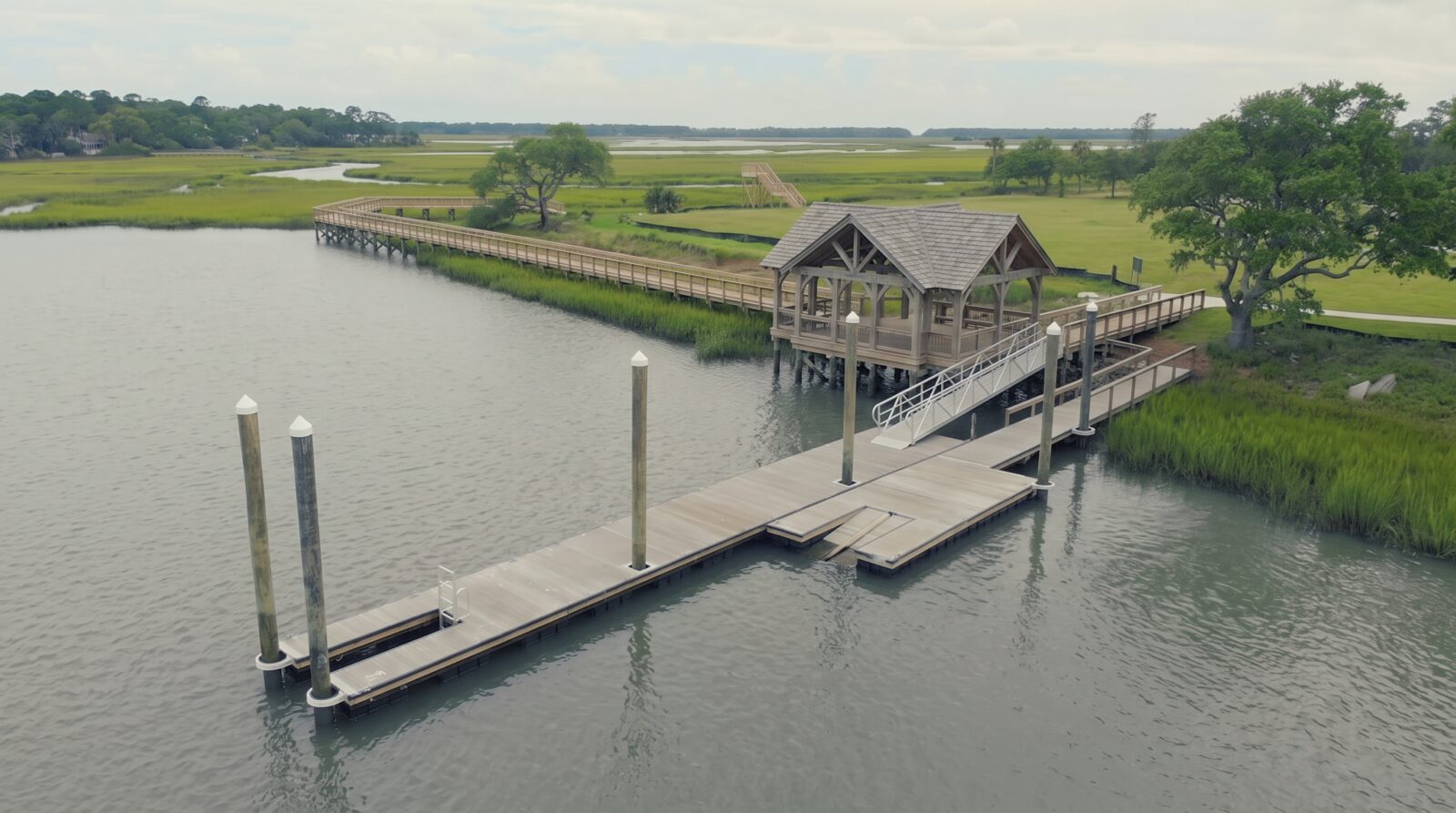 Aerial view of a shared community dock in a South Carolina waterfront neighborhood: a long timber walkway over spartina marsh leading to a covered pierhead, an aluminum gangway and a floating dock held by timber guide piles.
