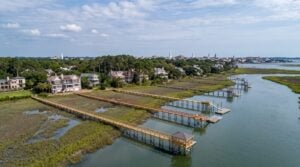 Aerial view of private timber docks with long marsh walkways and covered pierheads extending from a West Ashley shoreline, with the Charleston peninsula skyline and church steeples visible across the water.